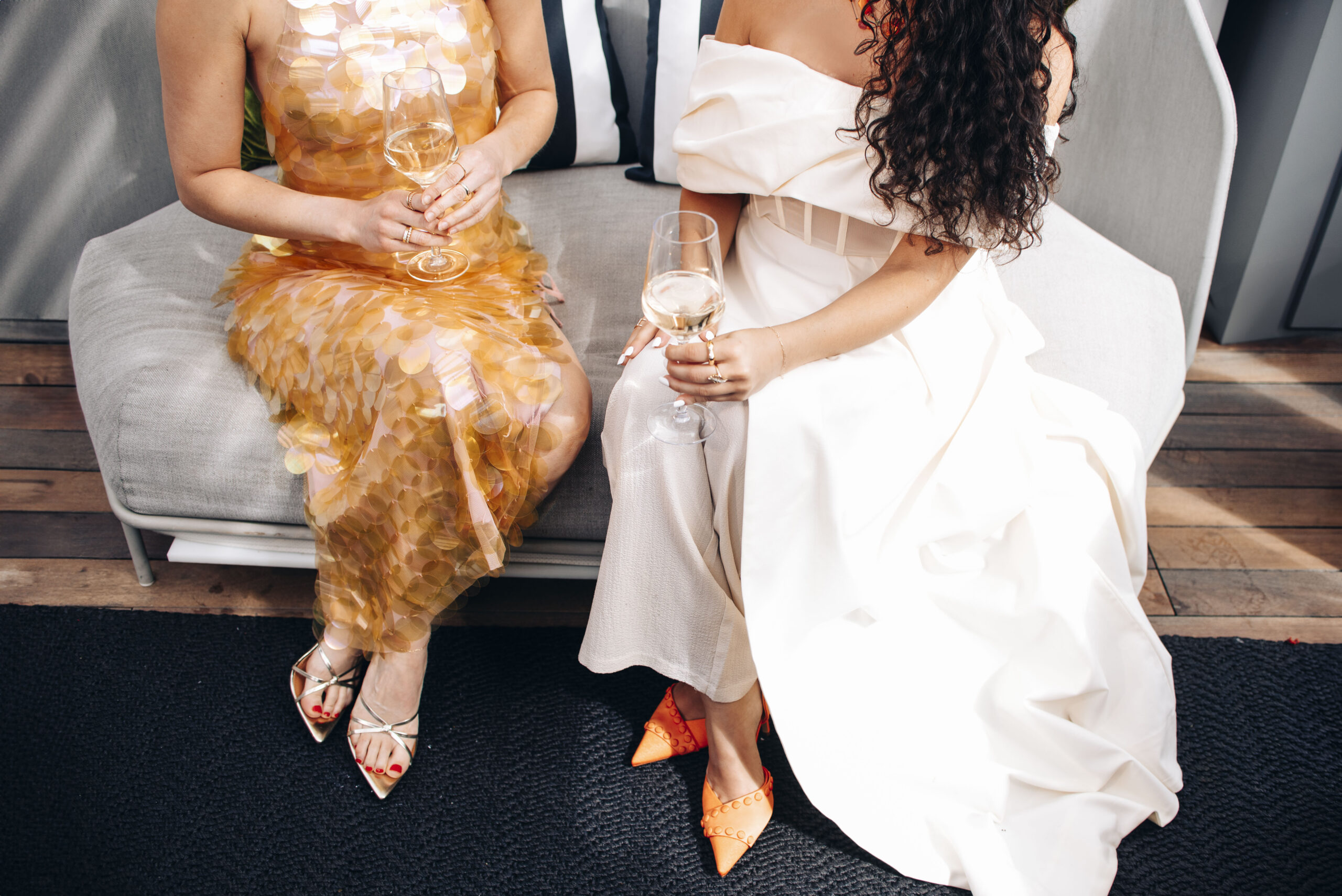 Two women talking on a couch in fashionable dresses and shoes while drinking wine.