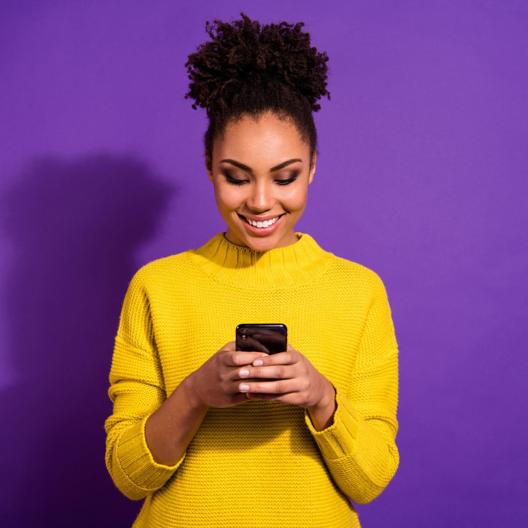 A woman in a yellow sweater standing in front of a purple wall. She's looking down at her cell phone.